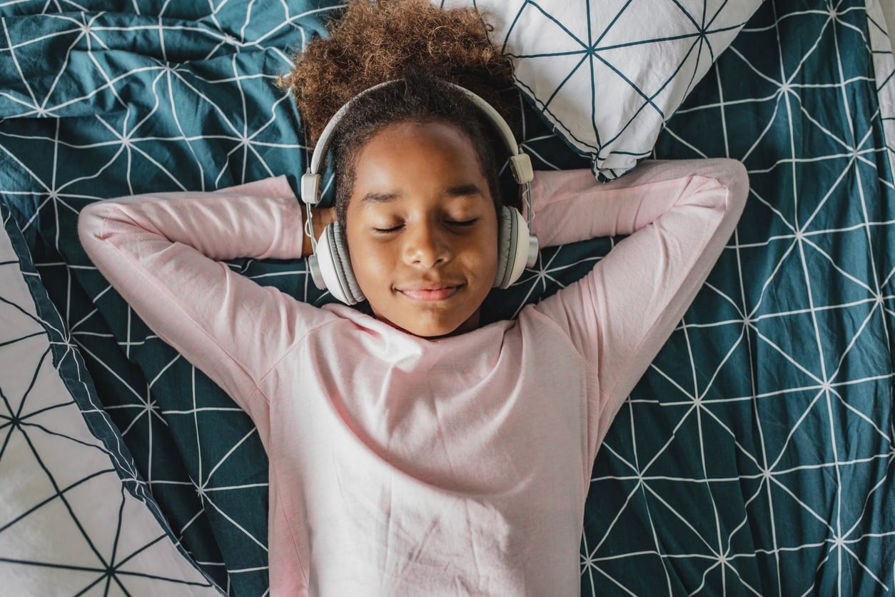A little African American girl enjoys her free time at home. Young girl laying in bed enjoying music, she is listening to music via wireless headphones