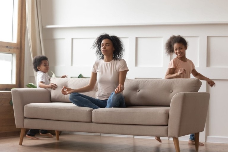 Calm african single mom doing yoga exercises sit on sofa reducing stress with active noisy kids playing at home, mindful black parent mother meditating relaxing with children running in living room