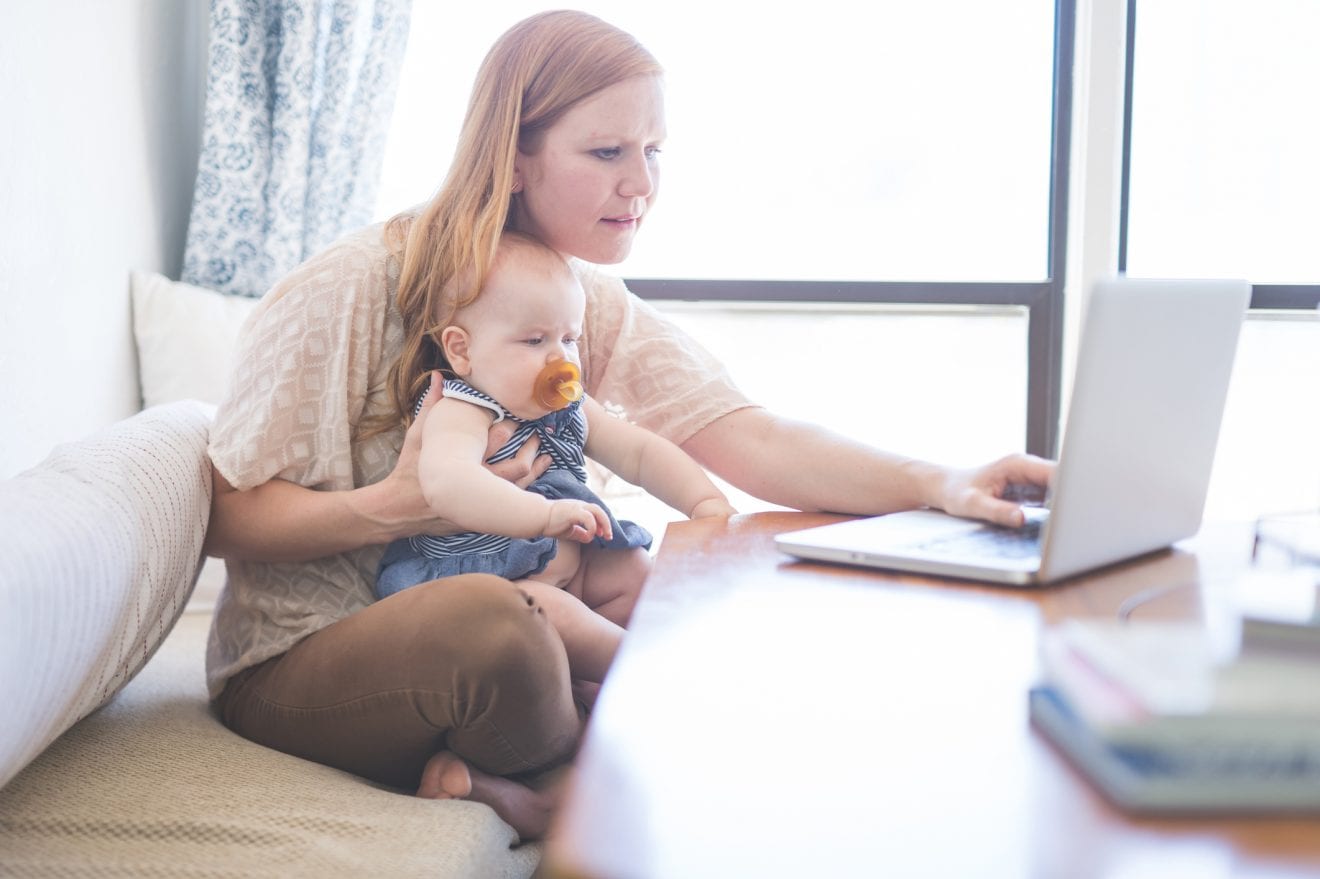 A young mom tries to simultaneously respond to emails on her laptop while holding her baby girl in her lap, who is staring at the screen with a pacifier in her mouth and trying to help mom out...