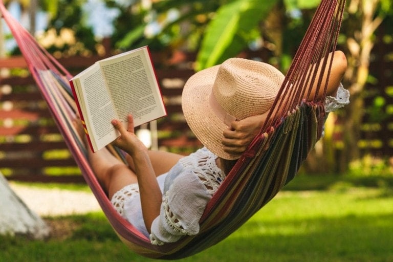 Happy woman in white dress relaxing in hammock reading a book.