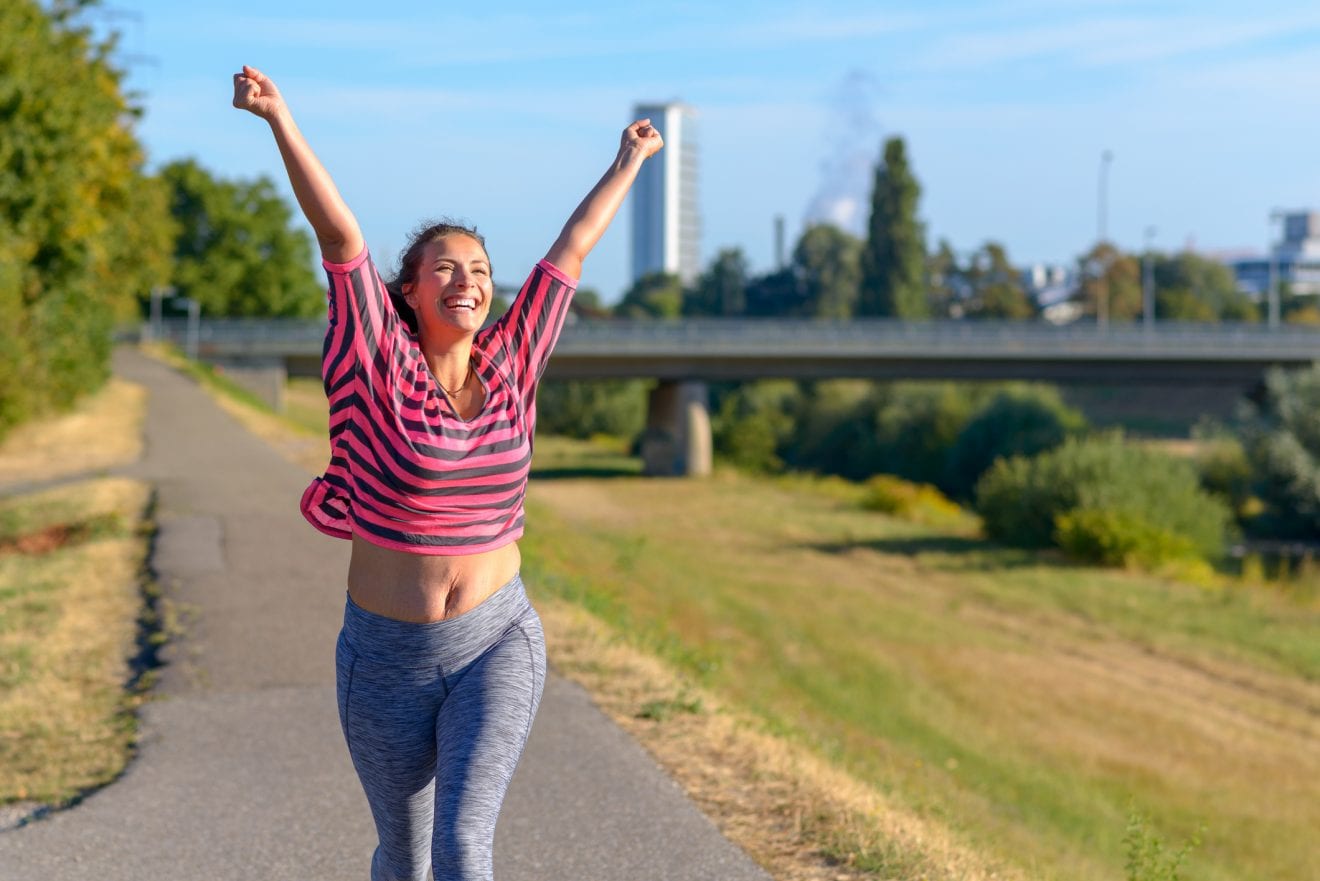 Happy fit young woman cheering and celebrating as she walks along a river after working out jogging