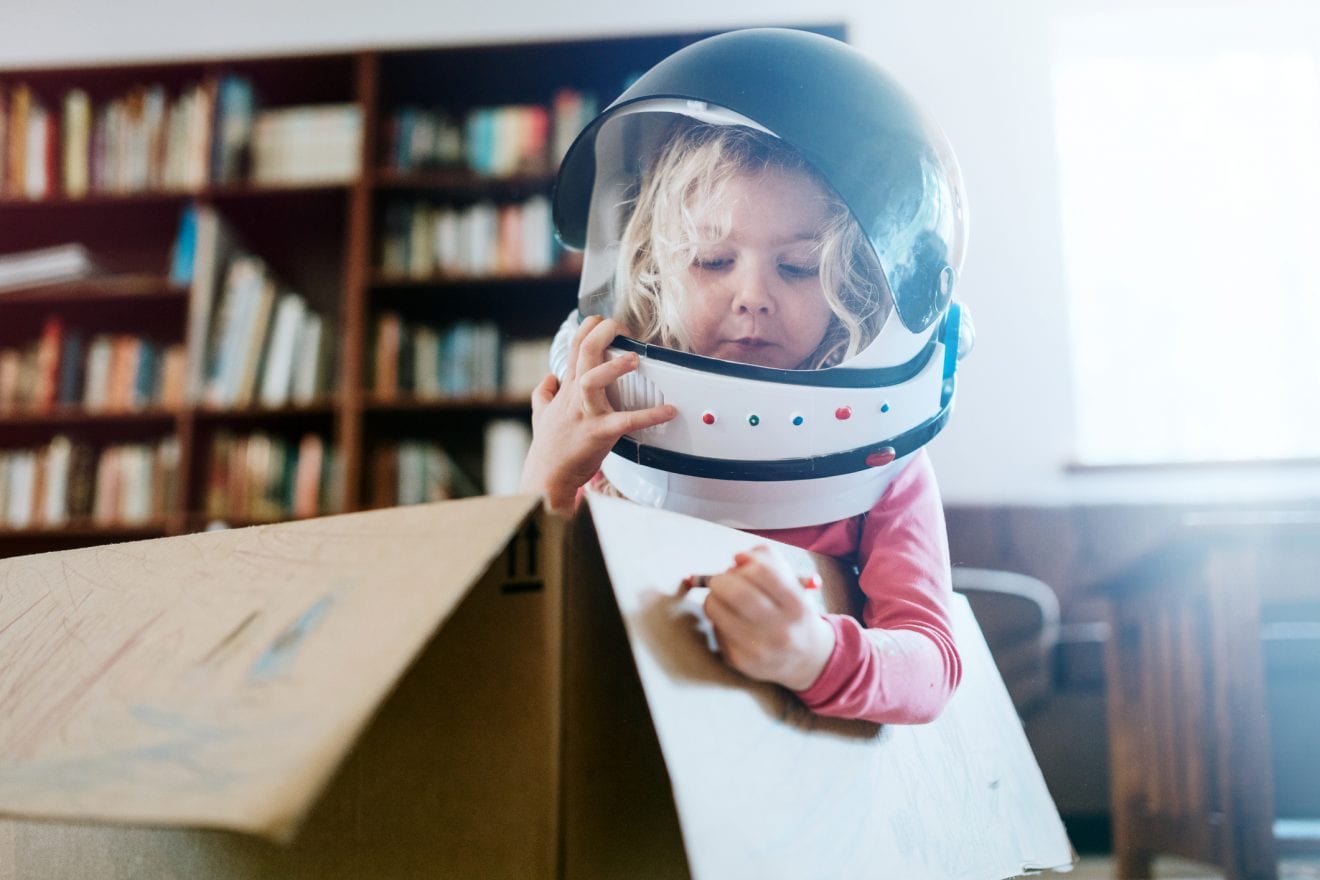 A boy and girl at home in their living room play in and color a cardboard box, one of them wearing a toy astronaut helmet. Imagination, discovery, and fun.