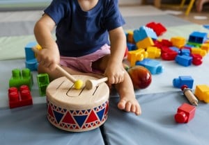 A gifted baby is playing with a drum using two drumsticks while sitting on a play mat. Various colorful building blocks are scattered around the child on the mat. The child is wearing a blue shirt and pink shorts.