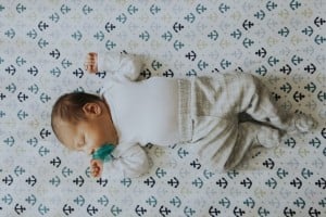 A baby, who needs to know about baby's sleep patterns, is lying on its back on a patterned sheet with anchors. The baby is wearing a white long-sleeve shirt, gray checkered pants, and has a pacifier in its mouth. The baby's left arm is raised while the right arm rests beside its body.