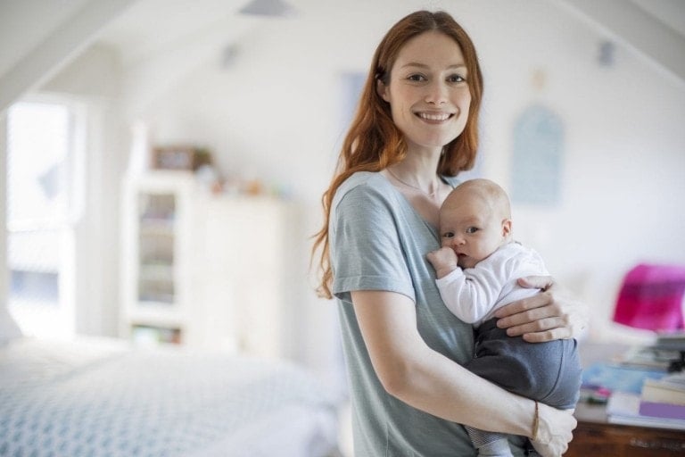 Smiling redhead woman standing with cute son. Portrait of beautiful mother is carrying newborn baby boy in bedroom. They are at home.