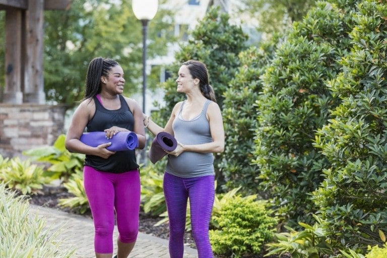 Two multi-ethnic pregnant women in their 30s, walking through a garden in the city, carrying yoga mats, conversing on their way to a yoga class for some exercise and relaxation.