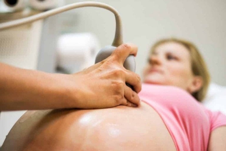 A woman in a pink top is lying down while a medical professional performs an ultrasound scan on her abdomen. The focus is on the ultrasound probe on her belly, making it crucial to choose or get a new OB-GYN for expert care.