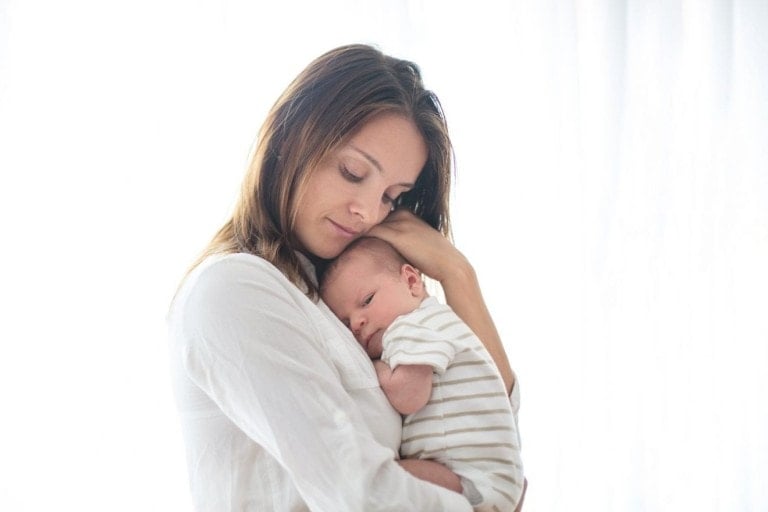 Young mother, holding her newborn baby boy at home in living room, back lit.