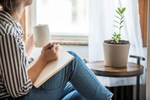 A person in a striped shirt and jeans is sitting by a window, holding a cup and writing in a notebook, fostering a positive mindset. A small potted plant is on a round wooden table next to them. Light filters through a white curtain, creating a serene atmosphere.