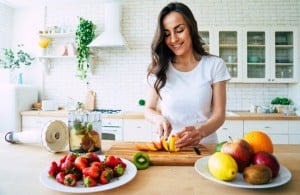A woman in a white t-shirt is slicing a peach on a wooden cutting board in a bright kitchen. Nearby are plates with a variety of fruits, including strawberries, kiwi, apples, and oranges. A blender filled with chopped fruits sits beside her, ready to create smoothies that boost your immune system.