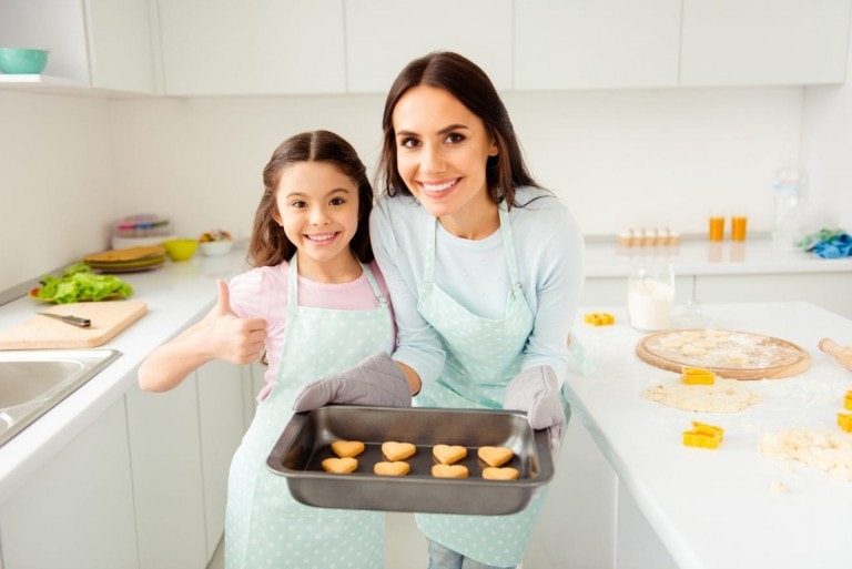 Charming adorable attractive beautiful brunette caucasian kind smiling young mum and her small little offspring daughter, wearing apron, showing thumb up, holding prepared cookies in light kitchen.