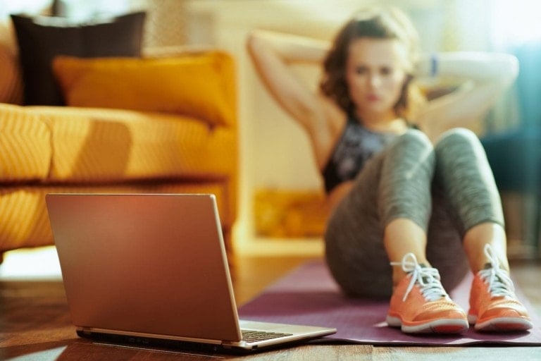 Closeup on beige laptop and woman in background doing abdominal crunches on fitness mat while watching fitness tutorial on internet in the modern house.