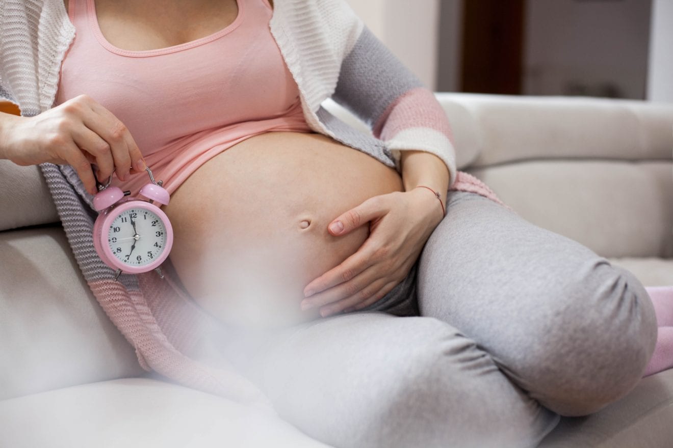 Pregnant woman holds hands on belly and looking on an alarm clock.