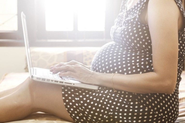 Sitting on the edge of a bed, a pregnant woman is typing on the keyboard of a laptop working on her baby registry.