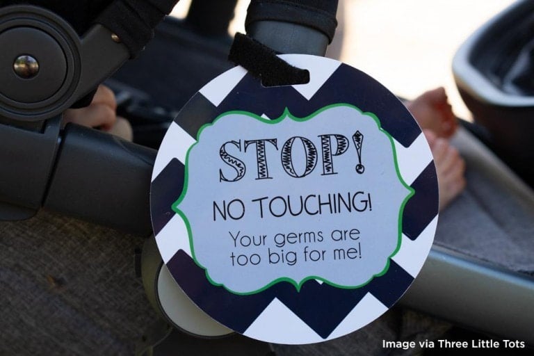 A round sign with a black and white zigzag pattern and green border hangs on a piece of baby equipment. The sign reads, "STOP! NO TOUCHING! Your germs are too big for me!" Partial view of a baby's foot is visible in the background, subtly asking people not to touch your baby.