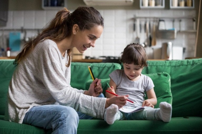 A woman and a child are sitting on a green sofa. The woman, presumably the babysitter, is holding a pencil and smiling at the child's drawing, while the child uses a red crayon on the paper. The background includes a kitchen area with various utensils, but there are no red flags in sight.