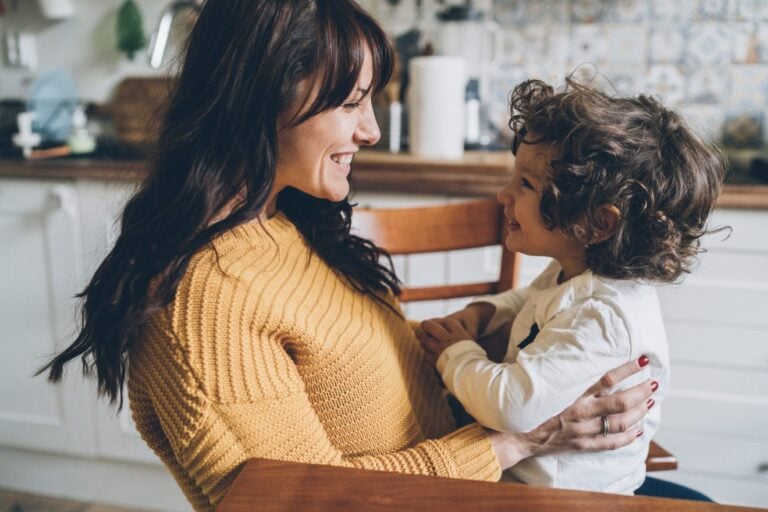 A mom in a yellow sweater sits in a kitchen, holding her toddler with curly hair who is sitting on a wooden chair. They are facing and smiling at each other. The background shows kitchen counters and tiled walls.
