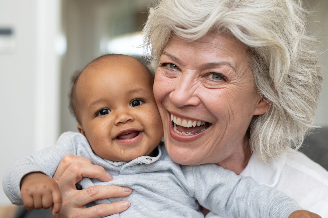 A Caucasian senior grandmother is fully present and spending time with her young, ethnic grandson at home. They are smiling and happy while sitting on a couch in this portrait.