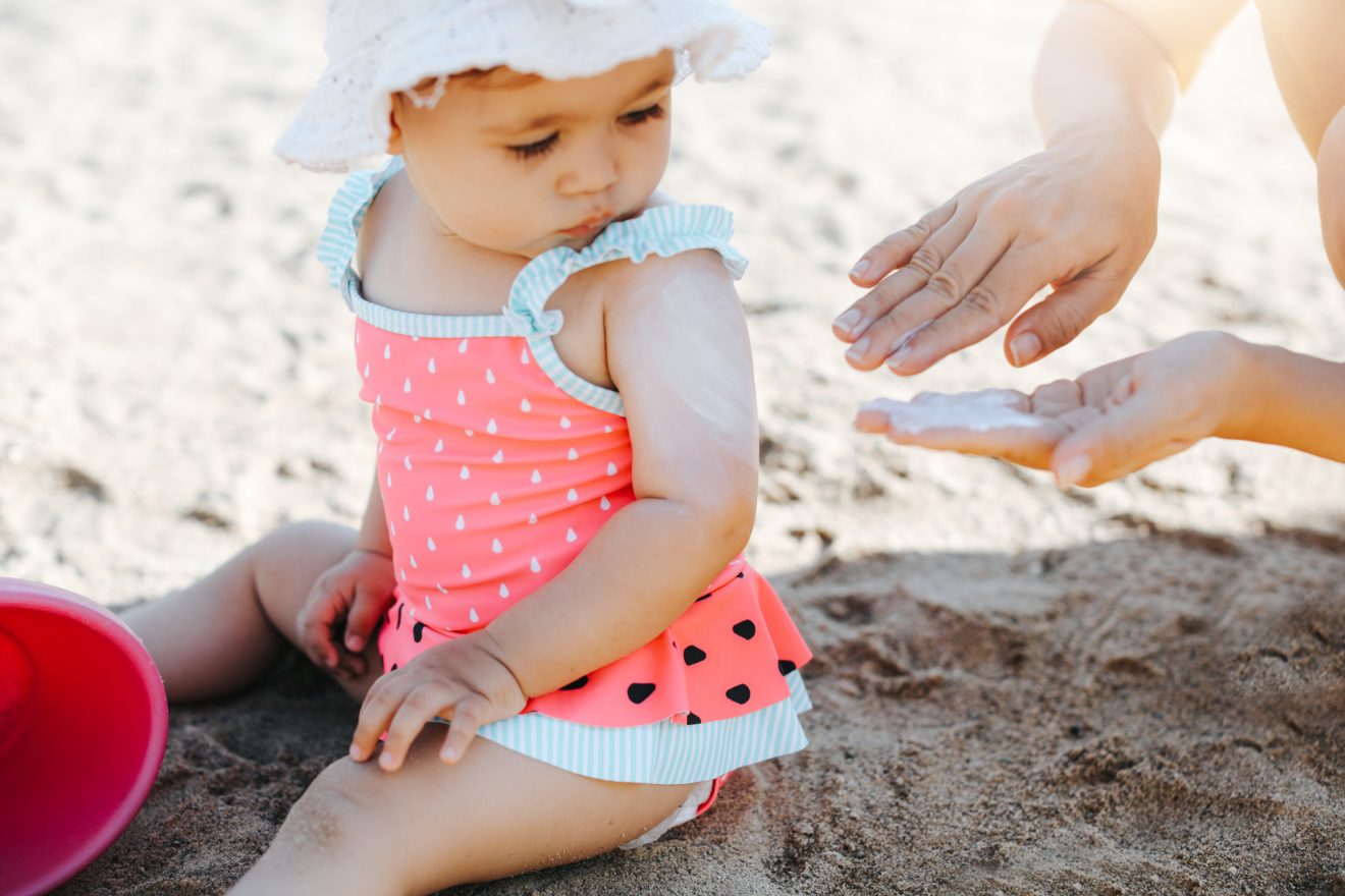 Toddler sitting on the beach in her swimsuit and hat with her parent rubbing sunscreen on their hands preparing to rub it on her.