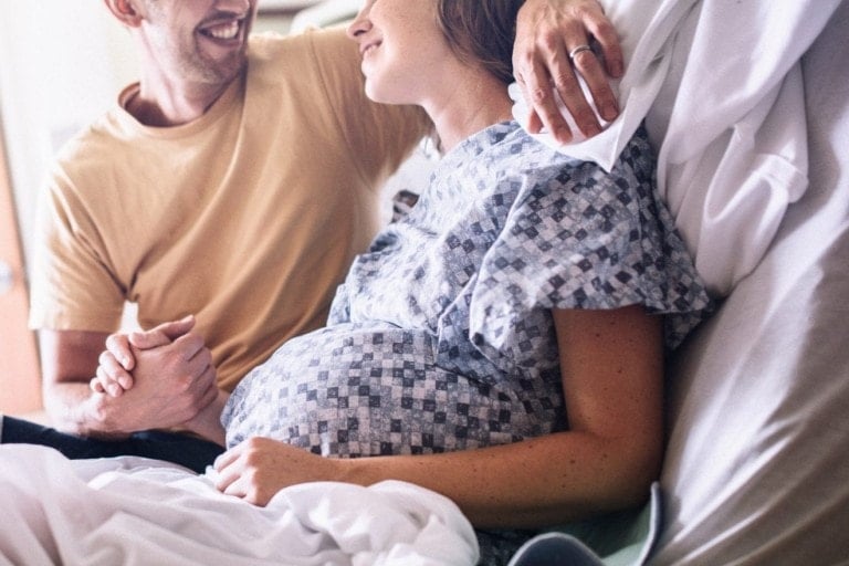 A smiling, happy woman in labor prepares to give birth in a clean white hospital setting. She holds the hand of her partner or husband as they look lovingly at each other. A depiction of a positive pregnancy and delivery.