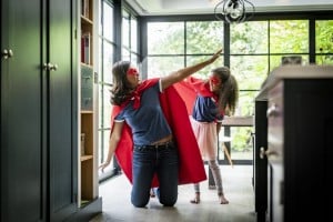 Playful mother and daughter in superhero costume against window. Woman is kneeling with arms outstretched by girl standing in kitchen.