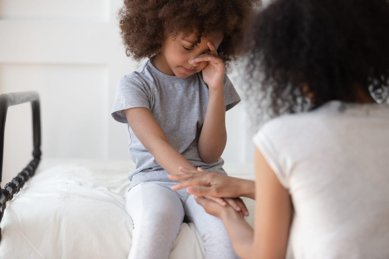 Mixed race mommy holding hand of upset little cute girl sitting on bed, wiping tears. African american worried woman talking, supporting, comforting small daughter, suffering from bullying.