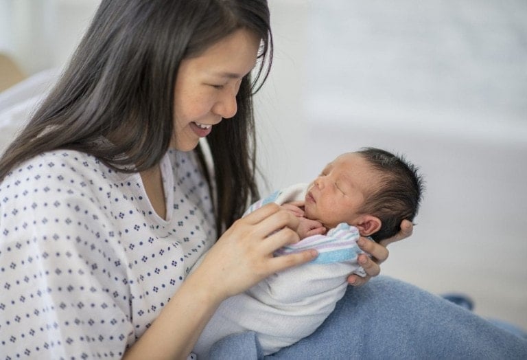 A mother and newborn baby boy are indoors in a hospital room. The mother is laying in bed and smiling down at her baby in her arms. The baby is sleeping.