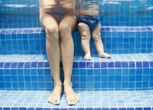 An underwater view of an adult and a baby sitting side by side on pool steps. The adult is wearing a striped swimsuit, while the baby is in a blue swim diaper. Both have their legs submerged, which slightly distorts the image. The pool steps are tiled in blue, creating a safe environment to prevent drowning.