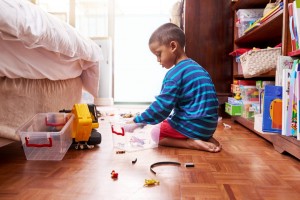 A child wearing a blue and black striped sweater and red shorts is sitting on a wooden floor, playing with toys next to a bed. There is a clear plastic container and a toy truck on the floor, with various toys scattered around. Shelves with more toys are in the background, perfect for teaching your child to pick up after themselves.