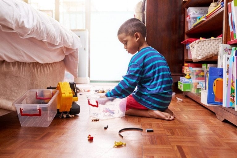 A young boy sitting on the floor picking up his toys in a bedroom.