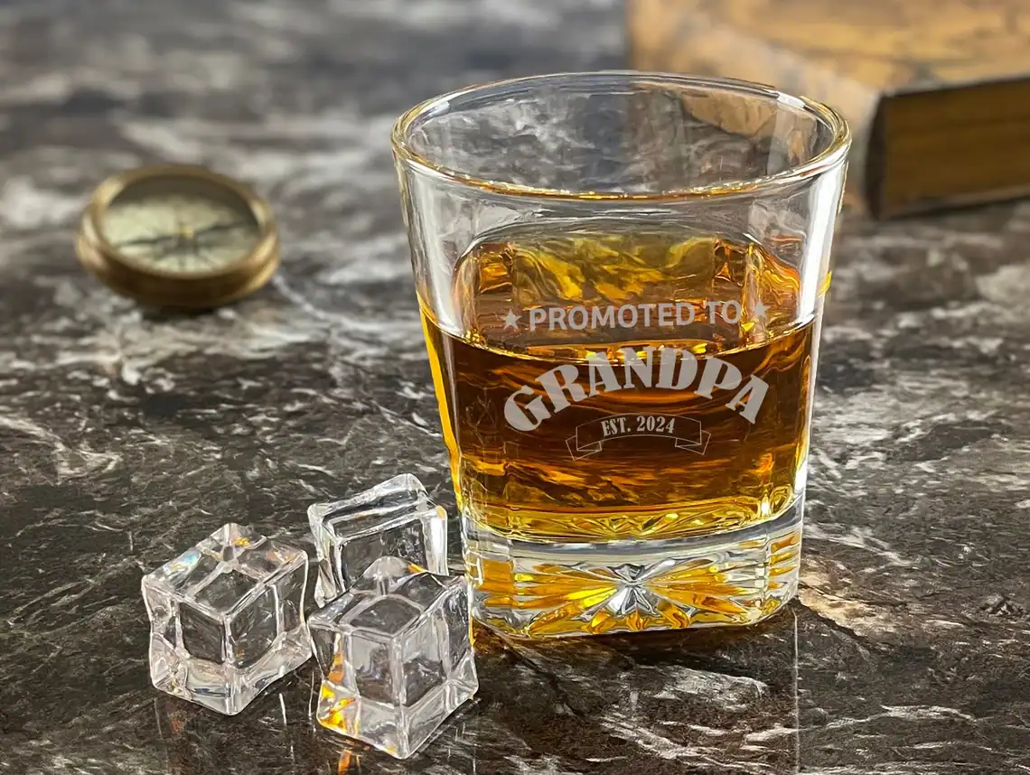 Whiskey glass with ice cubes and personalized "Grandpa" engraving on a rustic tabletop.