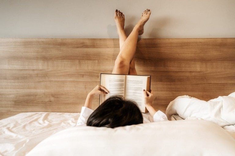 Asian woman lying down, legs crossed, she is reading a book on the bed