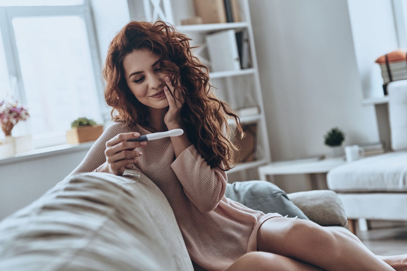 Attractive young women looking at pregnancy test and smiling while sitting on the sofa at home