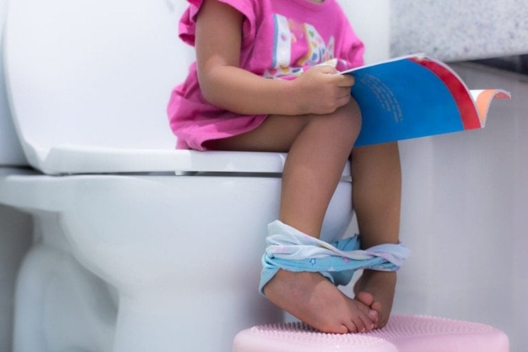 A young little girl sitting on a big toilet in the bathroom reading a book while waiting to go potty. Close up on legs.
