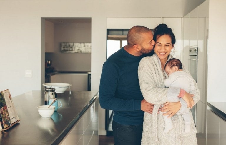 Man kissing his wife holding a newborn baby boy in kitchen.