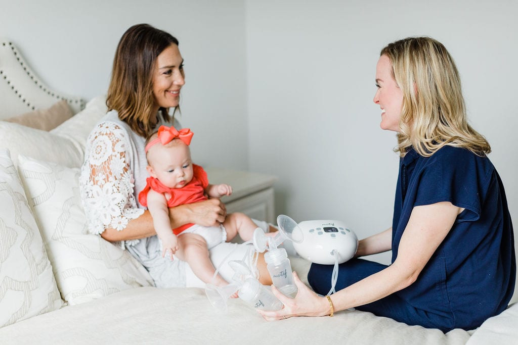 A postpartum doula sitting on the bed with her new mom client helping her with breast pump tips.