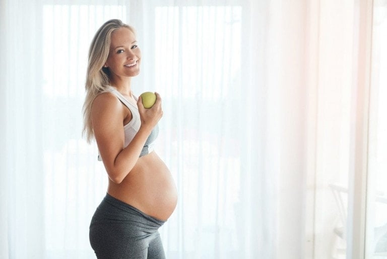 Portrait of a happy pregnant woman dressed in sportswear and eating an apple at home.