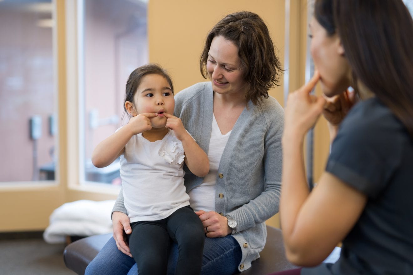 A speech therapist of Asian descent works with a young girl on pronunciation. They are sitting on a treatment table with their forefingers pointing to the sides of their mouths and practicing the correct way of forming sounds and syllables. The girl is sitting on her mom's lap.