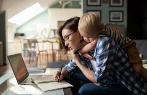 Side view of busy female in casual clothing studying with laptop and writing notes with little boy is hugging her while standing behind her on sofa in the living room.