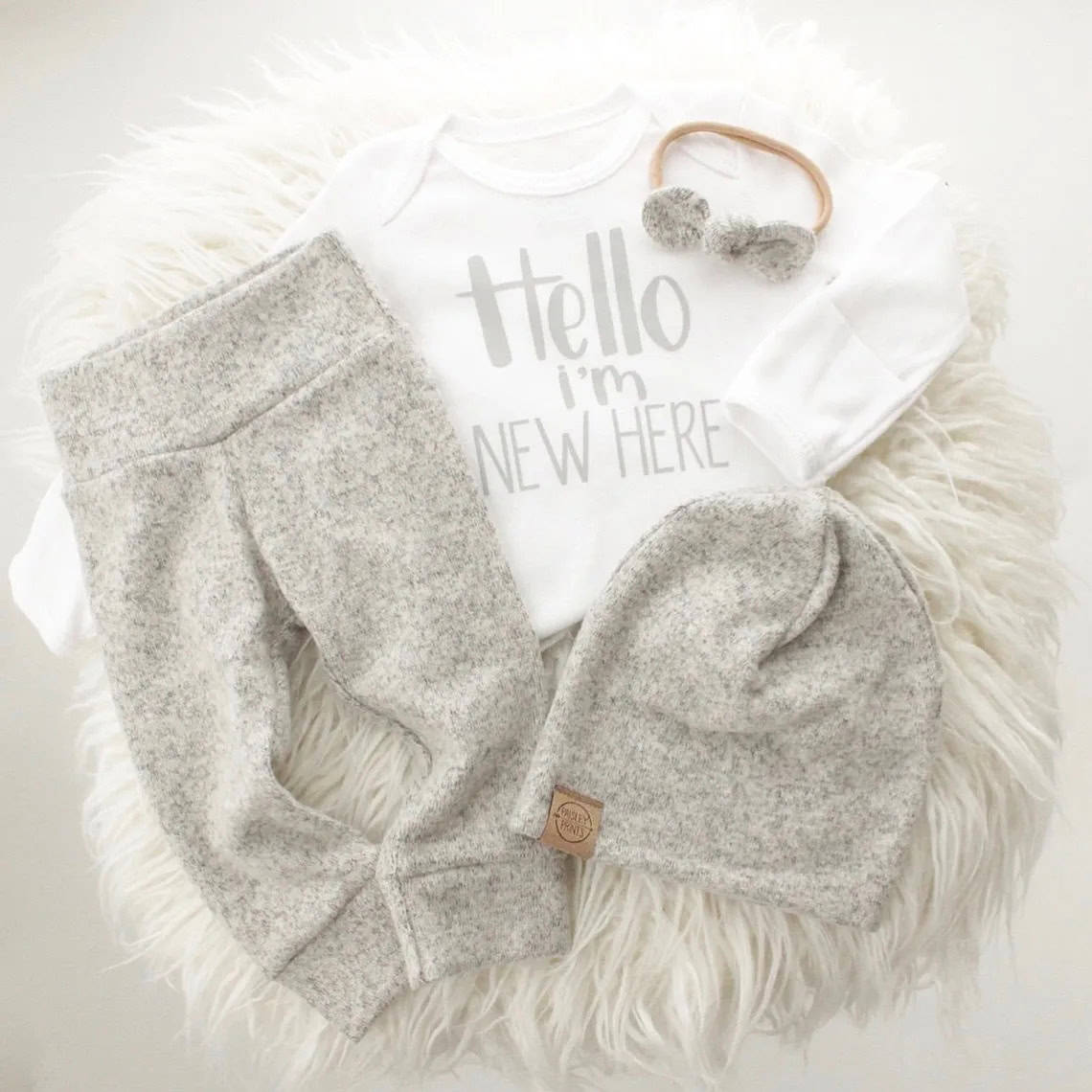 A baby coming home outfit is laid out on a soft white surface. It includes a white onesie with Hello Im new here printed in gray, gray pants, a matching gray beanie, and a gray headband with a bow.