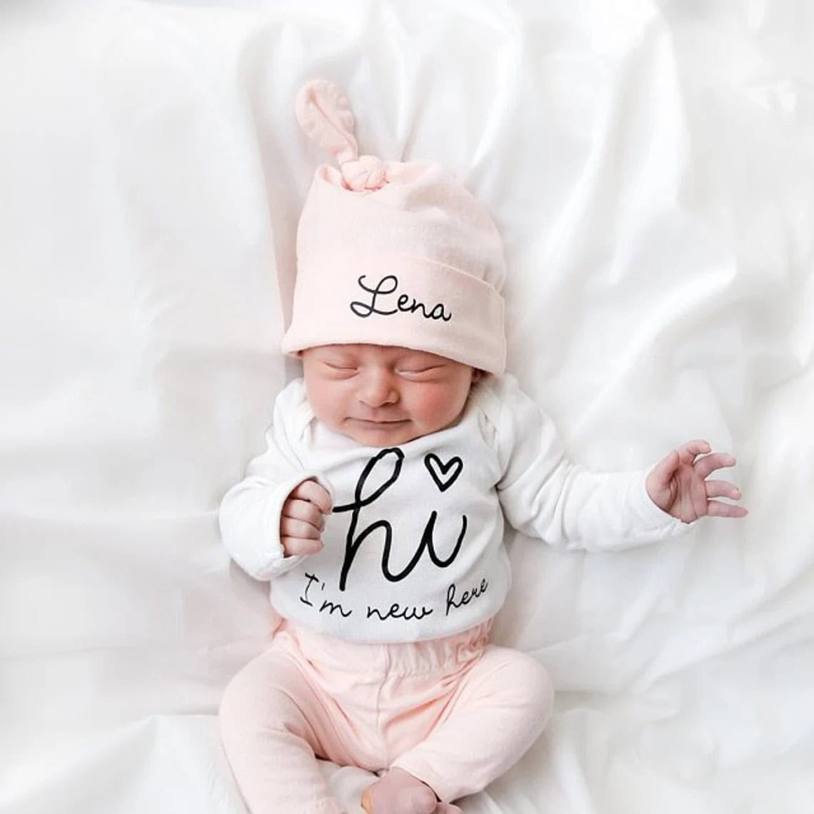 A newborn baby with eyes closed, wearing a pink hat labeled Lena and a cozy coming home outfit that says hi, I'm new here, is lying on a soft white surface.