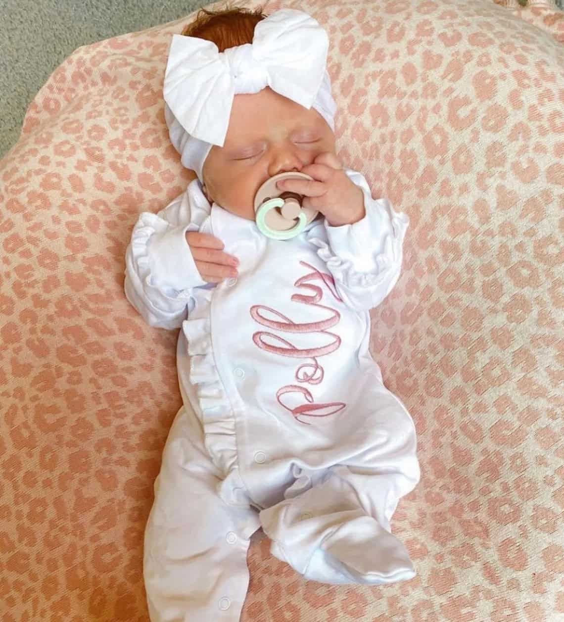 A baby is lying on a pink and white patterned blanket, perfect for her coming home outfit. She wears a white ensemble with ruffled sleeves, a matching bow headband, and clutches a pacifier. The name Polly is sweetly written in pink on her adorable attire.