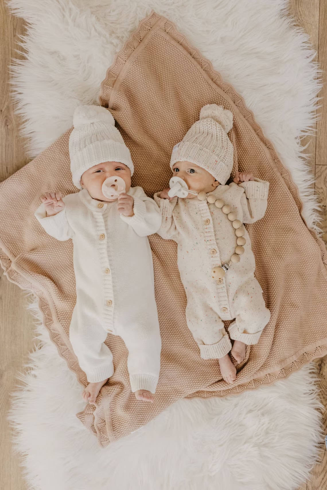 Two babies are lying on a soft, textured blanket atop a fluffy rug. Both wear knitted white onesies and matching beanies—perfect coming home outfits. Each baby has a pacifier, and one holds a beaded pacifier clip. The scene is cozy and neutral-toned.