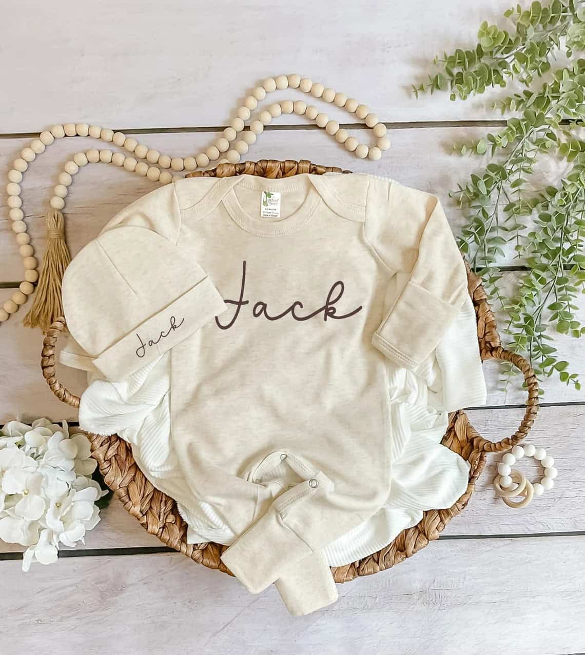 A babys beige coming home outfit, adorned with the name Jack, rests in a woven basket. Its beautifully surrounded by wooden beads, greenery, and delicate white flowers on a light wooden surface.
