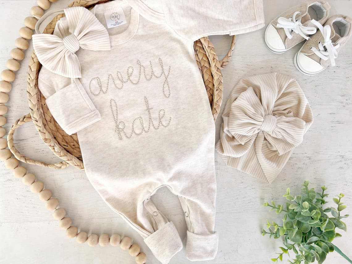 A charming display of coming home outfits features a neutral-toned baby set, including a romper with avery kate stitched on it, paired with a matching headband, hat, and shoes. A wooden beaded garland and small green plant complete the scene on a light-colored woven surface.