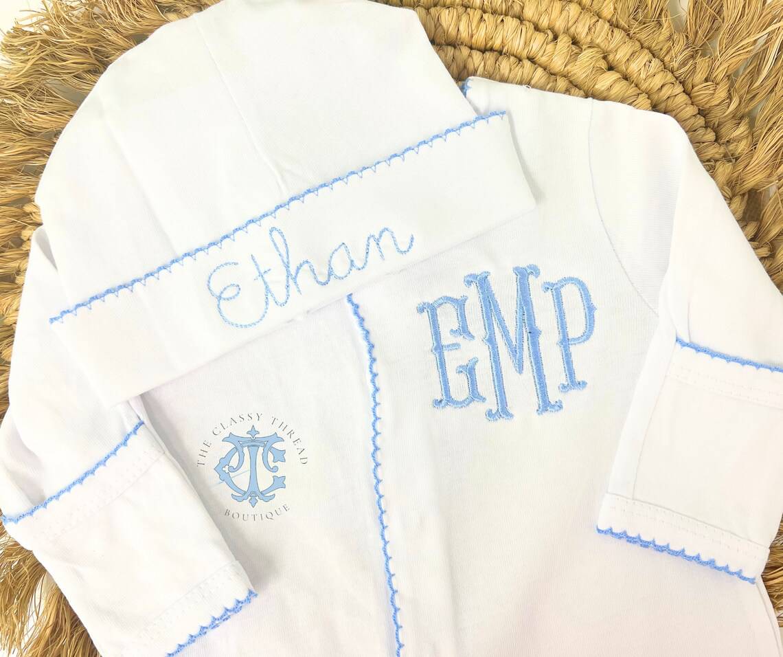 A baby coming home outfit and hat are displayed on a woven surface. The white ensemble features light blue embroidery with the initials EMP and the name Ethan adorning the hat. The logo The Classy Thread Boutique is elegantly visible, promising style from your first day home.