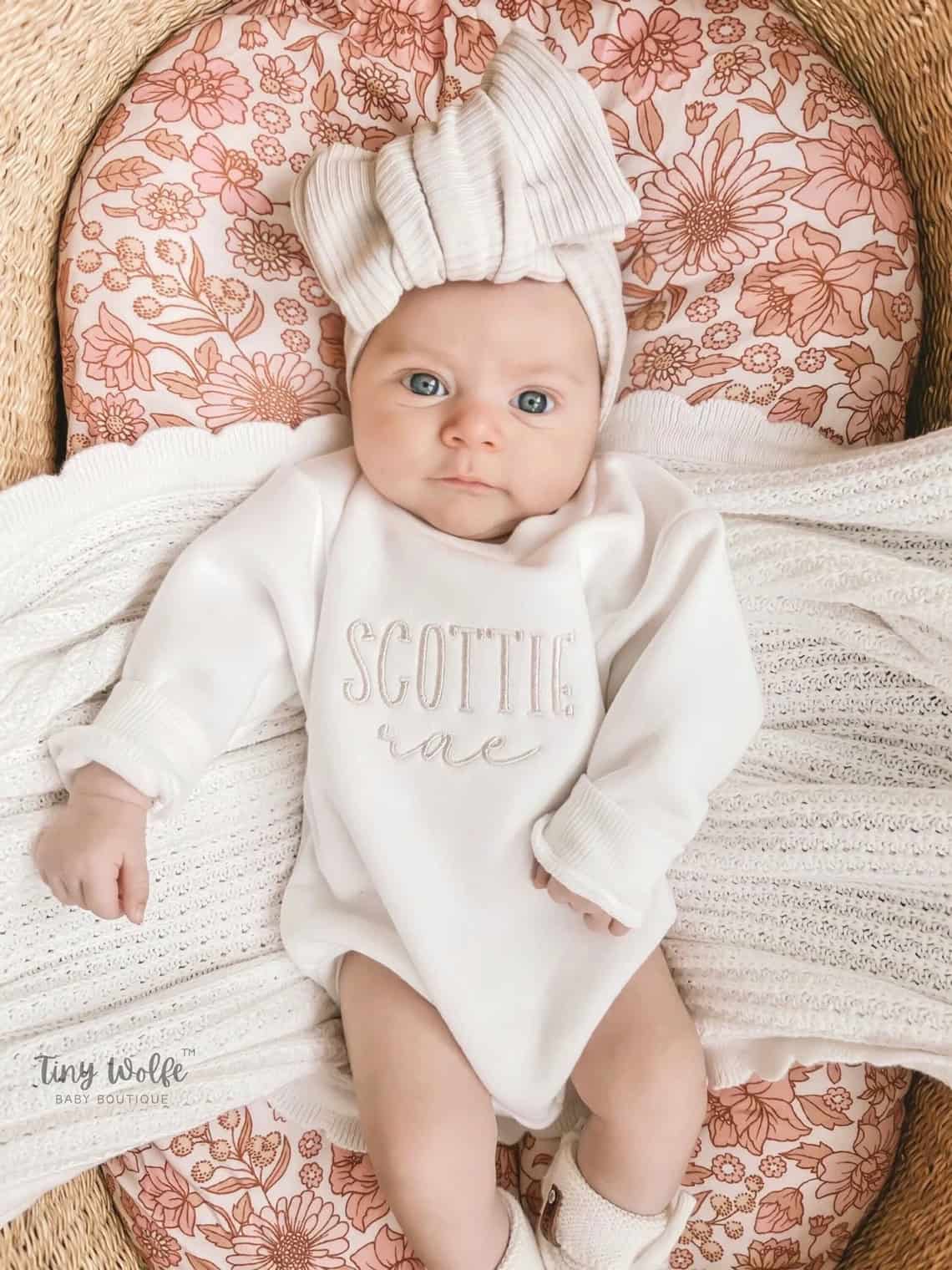 A baby in a charming coming home outfit with Scottie Rae embroidered on the front lies on a floral-patterned cushion. Adorned with a large bow headband and wrapped in a knit blanket, the little one gazes at the camera with wide-open eyes.