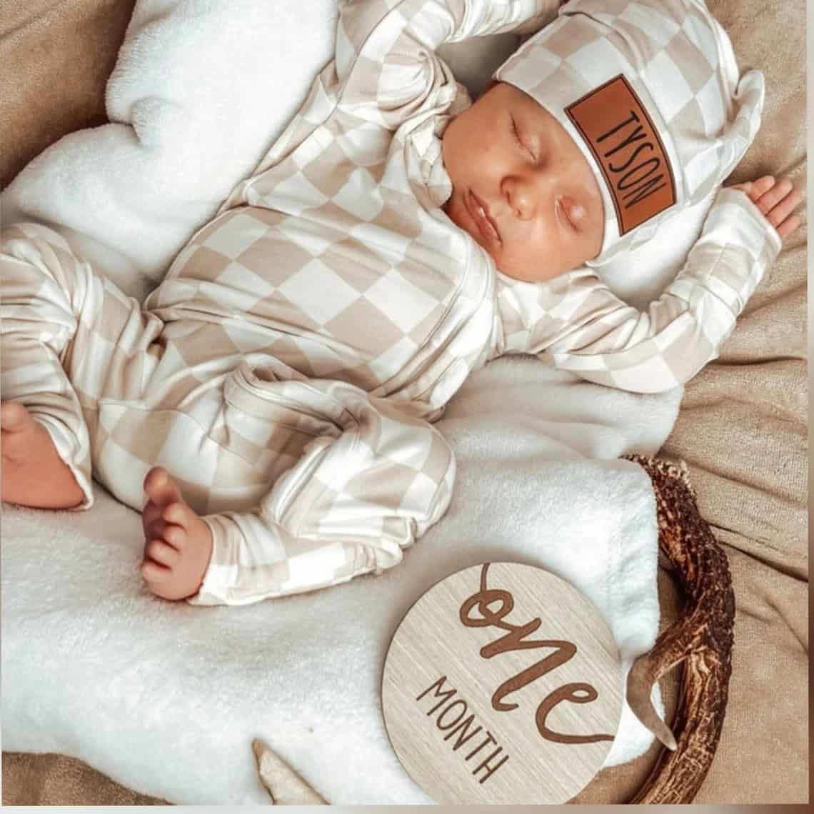 A baby, cozy in a beige and white checkered coming home outfit and hat, sleeps on a soft white blanket. A wooden sign beside the peaceful little one reads one month, as the baby rests serenely with tiny arms raised, marking their first milestone in style.