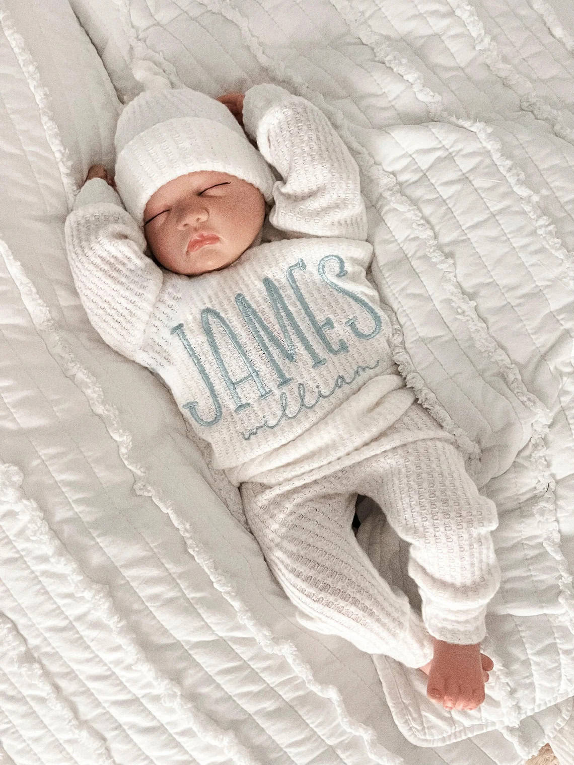A baby in a white knit coming home outfit and hat sleeps peacefully on a quilted blanket. James is embroidered in blue across the chest. The little one rests on their back, arms gently raised above their head.