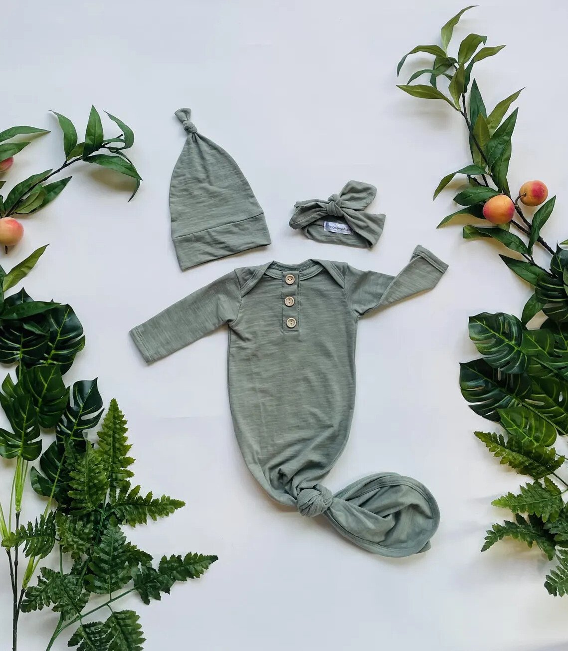 A green infant knotted gown, ideal for coming home outfits, features buttons and is paired with a matching hat and headband. The set is elegantly displayed on a white background, surrounded by green leaves and small, round fruits.